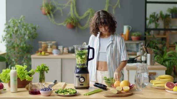 Junge Frau gibt frische Salatblätter in den Mixer, während sie zu Hause in der Küche grünen Smoothie kocht - Filmmaterial, Video