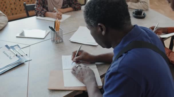 Captura selectiva de enfoque del hombre negro senior haciendo tarea de escritura durante la lección de inglés para estudiantes inmigrantes - Imágenes, Vídeo