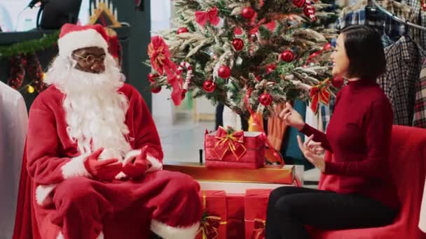 Азиатская женщина, участвующая в разговоре с сотрудником, выдающим себя за Санта Клауса во время праздничного рождественского шопинга. Подарок на Рождество от работника модного магазина - Кадры, видео