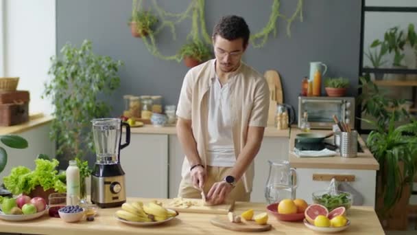 Medio largo tiro de hombre joven cortando plátano en la mesa de la cocina mientras se preparan ingredientes para hacer batido saludable en casa - Imágenes, Vídeo