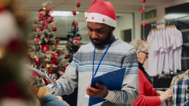Funcionário afro-americano navegando através de prateleiras de roupas na loja de moda decorada de Natal, escrevendo os preços ajustados na área de transferência. Trabalhador inspecionando blazers, à procura de danos - Filmagem, Vídeo