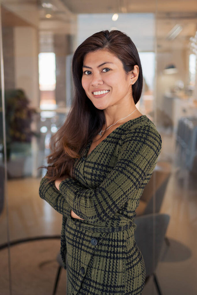 Zijaanzicht van positieve jonge Aziatische vrouw in elegante jurk glimlachen en kijken naar camera terwijl staande met de armen gekruist in het kantoor - Foto, afbeelding