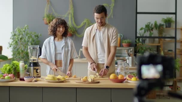 Επιλεκτική φωτογραφία εστίασης του νεαρού ζευγαριού να κόβει φρέσκα φρούτα και να μιλάει ενώ τραβάει υγιεινά vegan food vlog με ψηφιακή κάμερα στην κουζίνα στο σπίτι - Πλάνα, βίντεο