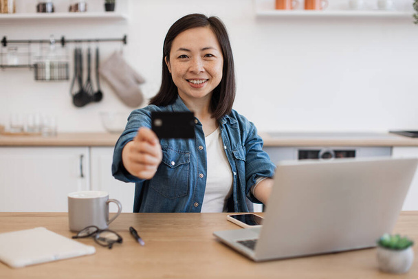 Focus on smiling asian lady reaching out credit card while sitting at kitchen table with gadgets on surface. Cheerful freelancer promoting cashless transactions via home banking service for customers. - Photo, image