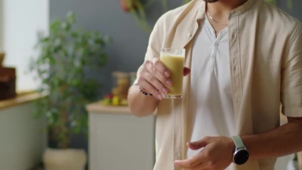 Tilt up πλάνο του νεαρού μεσανατολίτη άνδρα πίνοντας φρέσκα φρούτα smoothie από γυαλί, ενώ στέκεται στην κουζίνα στο σπίτι - Πλάνα, βίντεο