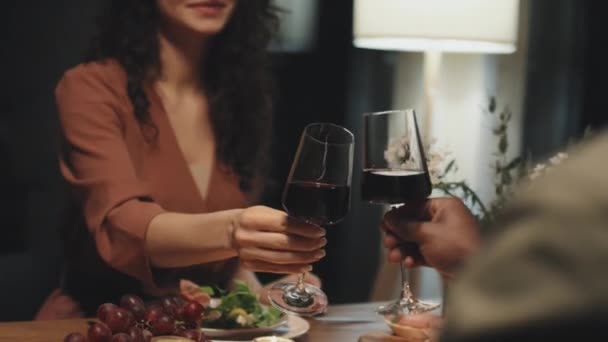 Жорсткий середній знімок кавказької жінки в червоній мідній блузці смердить келихами вина з чорним чоловіком, посміхаючись, обидва сидять за обіднім столом вдома - Кадри, відео