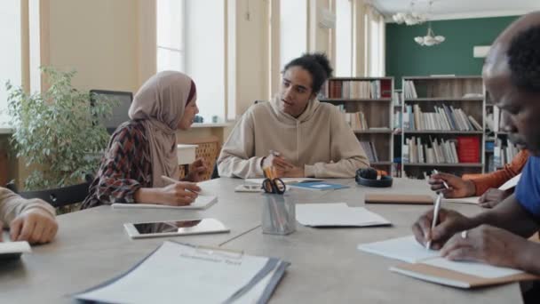 Joven hombre negro y mujer musulmana ayudándose mutuamente con la tarea de escribir durante la lección para los estudiantes inmigrantes - Metraje, vídeo