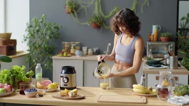 Longo tiro médio de jovem mulher magra em sutiã esportivo e leggins derramando smoothie de frutas do liquidificador em vidro e bebendo-o na cozinha em casa - Filmagem, Vídeo