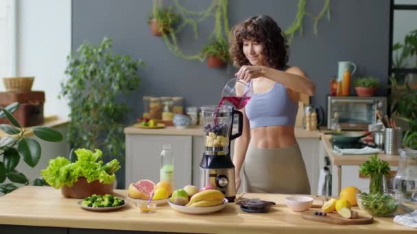 自宅でキッチンで健康的なスムージーを作っている間,ベリージュースをブレンダーに注ぐスポーツウェアの若いフィット女性の中長いショット - 映像、動画