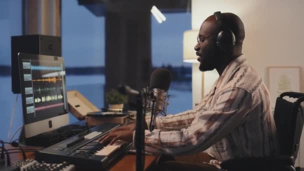 Strak medium shot van Afro-Amerikaanse man opnemen van audio tijdens het dragen van een koptelefoon, spelen toetsenbord en zingen in de microfoon, zijaanzicht - Video