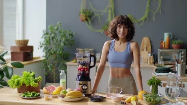 Retrato longo médio de mulher jovem em sportswear em pé na mesa da cozinha com frutas frescas e liquidificador e posando para câmera com sorriso - Filmagem, Vídeo