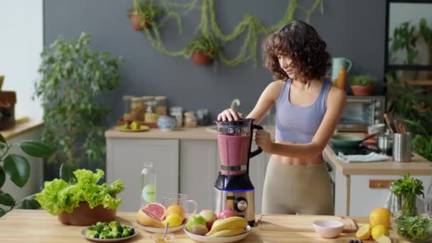 Bogenaufnahme einer jungen, fitten Frau in Sportkleidung, die zu Hause in der Küche Obst- und Beeren-Smoothie im Mixer zubereitet - Filmmaterial, Video