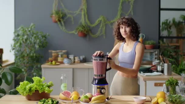 Portrait of beautiful slim girl in sports bra and leggings making smoothie in blender and smiling on camera while posing in kitchen at home - Footage, Video