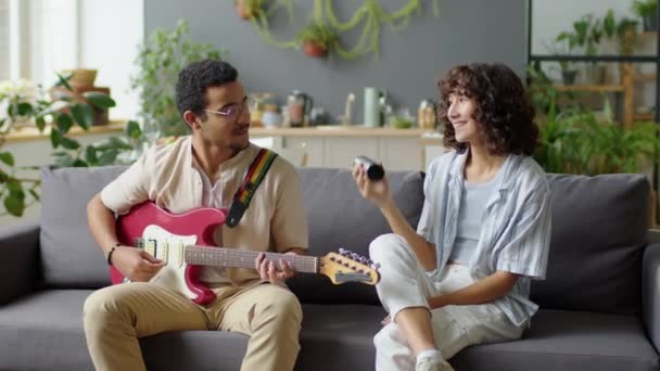 Fiatal gen Z pár ül kanapén a nappaliban, gitároznak és rázzák és élvezik a zenét, miközben együtt töltik a napot otthon - Felvétel, videó