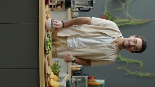 Retrato vertical de hombre vegano gen Z sonriendo y posando para la cámara en la mesa de la cocina con frutas frescas, verduras y licuadora en ella - Metraje, vídeo