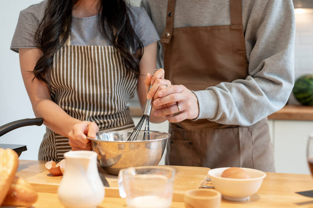 Nahaufnahme eines schönen und glücklichen jungen asiatischen Paares in einer Schürze, das Ei und Mehl miteinander verquirlt, zu Hause ein Kochdatum hat und in der Küche Pfannkuchen backt. - Foto, Bild