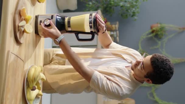 Retrato vertical tiro de gen Z homem vegan segurando liquidificador ao fazer smoothie e posando para câmera com sorriso na cozinha em casa - Filmagem, Vídeo
