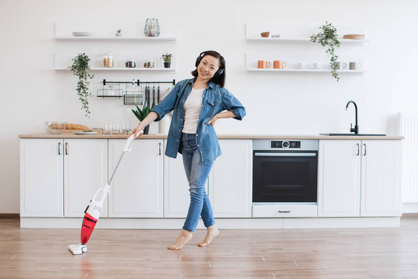 Cheerful woman vacuuming floor with cordless machine while listening energetic music in wireless headphones. Active asian female keeping house clean with good mood. Background of spacious kitchen. - Photo, Image
