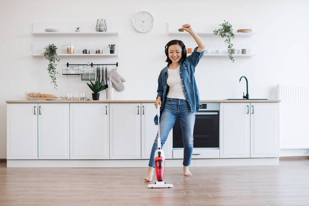 Smiling vietnamese woman wiggling with uplifting music in headphones while getting floor clean with cordless vacuum. Beautiful housewife entertaining with electric appliance while doing home care. - Фото, изображение
