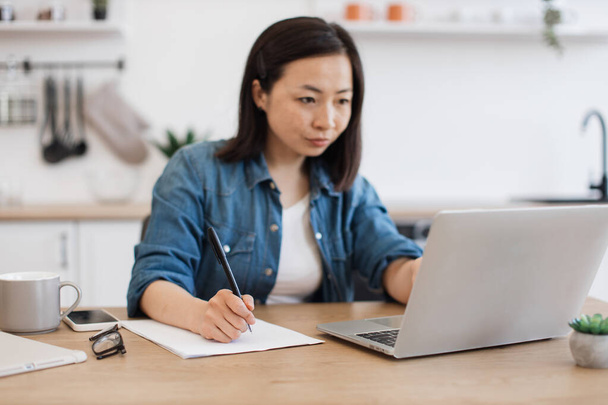 Side view of japanese lady in denim shirt putting down ideas on paper while looking through information on laptop. Thoughtful young woman planning e-commerce investment strategy in home office. - Fotoğraf, Görsel