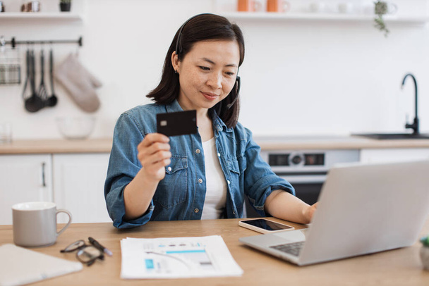Focused young female in headset holding black credit card while sitting behind computer screen in home interior. Relaxed asian employee doing online banking transaction using wireless connection. - Foto, imagen
