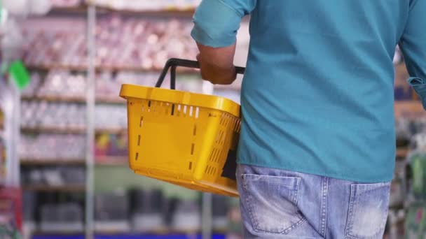 Vista trasera del comprador caminando en el pasillo del supermercado sosteniendo la cesta en la mano, primer plano detallado de la mano del consumidor en la tienda de comestibles - Imágenes, Vídeo