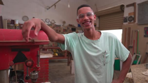 Retrato de um jovem trabalhador negro brasileiro sorrindo para a câmera ao lado da máquina industrial vestindo proteção ocular, expressão alegre feliz - Filmagem, Vídeo