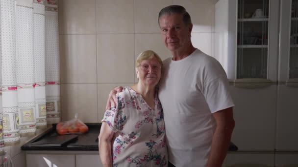 Oudere moeder en volwassen zoon van middelbare leeftijd poseren samen voor de camera, staande door keukenraam - Video