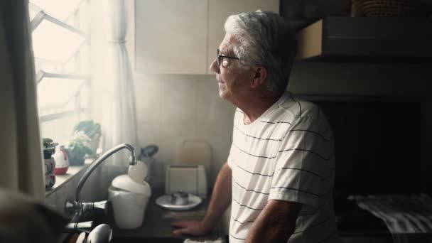 Pensive старший чоловік спирається на кухонний лічильник, дивлячись через вікно з продуманим виразом - Кадри, відео