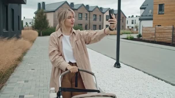 Captura de seguimiento de la joven madre moderna grabación de vídeo para el blog en el teléfono inteligente mientras camina al aire libre con su bebé en el cochecito - Metraje, vídeo