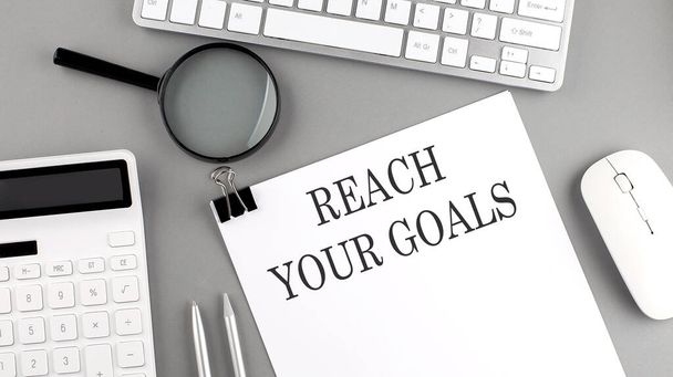 REACH YOUR GOALS auf Papier geschrieben mit Bürowerkzeugen und Tastatur auf grauem Hintergrund - Foto, Bild
