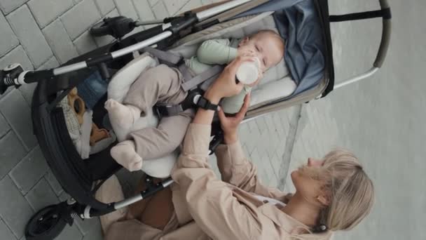 Tiro vertical de la joven madre biberón caucásica moderna alimentando a su bebé en el cochecito durante el paseo al aire libre - Imágenes, Vídeo