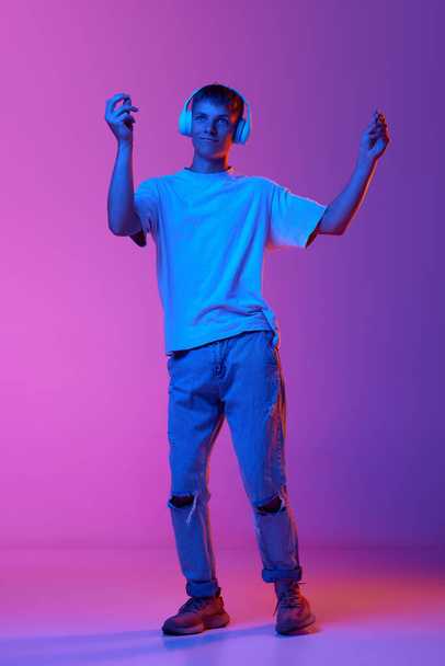 Full-length of young guy listening to music in headphones and dancing over gradient pink purple background in neon light. Concept of human emotions, youth, lifestyle, fashion, facial expressions, ad - Foto, afbeelding