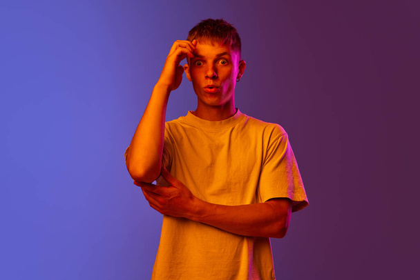 Young guy in white t-shirt standing with shocked face against gradient purple background in neon light. Concept of human emotions, youth, lifestyle, fashion, facial expressions, ad - Foto, Bild