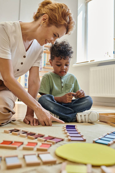 Escuela Montessori, profesor feliz señalando bloques de madera de colores cerca de chico afroamericano, inteligente - Foto, Imagen