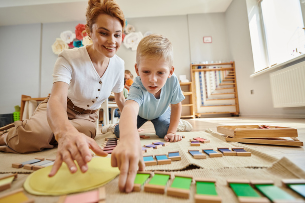 montessori escuela, rubia chico jugando juego educativo cerca feliz profesor, movimiento, juego de colores - Foto, imagen