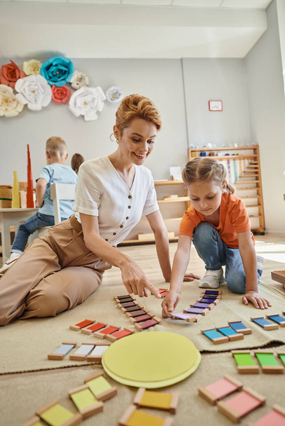 montessori szkoły, słodkie dziewczyny gra kolor dopasowania gry w pobliżu szczęśliwy nauczyciel kobieta, siedzi na podłodze - Zdjęcie, obraz