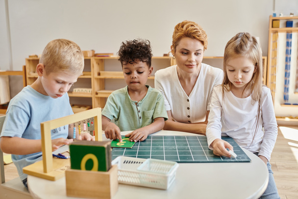 koncepcja szkoły montessori, wielokulturowe dzieci, pisanie dziewczyn na tablicy w pobliżu nauczycieli i chłopców - Zdjęcie, obraz