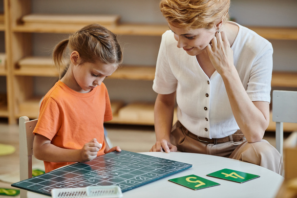 χαριτωμένο κορίτσι κοιτάζοντας chalkboard κοντά σε αριθμούς, μάθηση μέσω του παιχνιδιού, δάσκαλος, Montessori σχολείο - Φωτογραφία, εικόνα