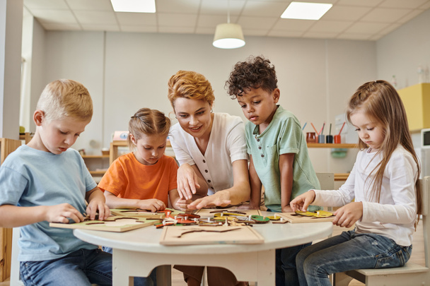 улыбающийся учитель играет с многонациональными детьми рядом с игрой на столе в школе Монтессори - Фото, изображение