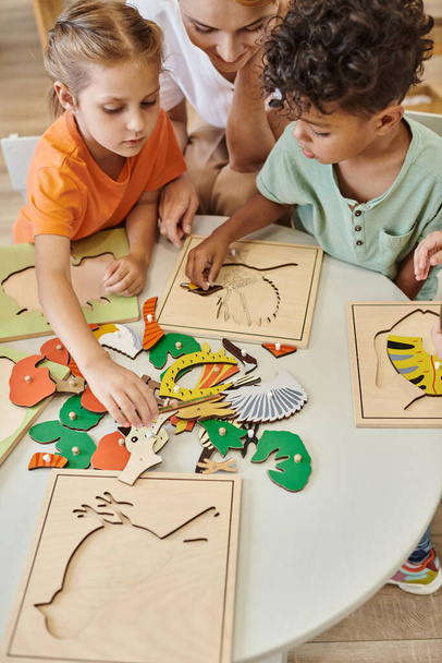 високий кут зору багатоетнічних дітей, які грають біля вчителя під час уроку в школі Монтессорі - Фото, зображення