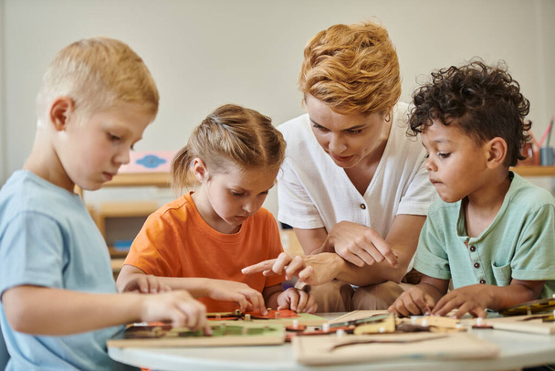 Lehrer im Gespräch mit multiethnischen Kindern in der Nähe didaktischer Materialien in der Montessori-Schule - Foto, Bild
