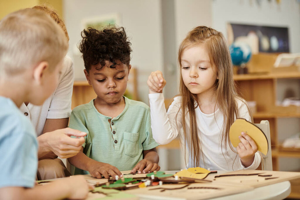 girl playing with didactic material near interracial kids and teacher in montessori school - Photo, Image