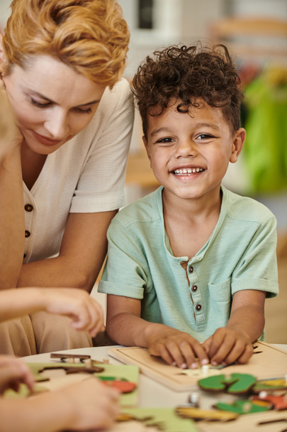 cheerful african american boy playing near teacher and looking at camera in montessori school - Photo, Image