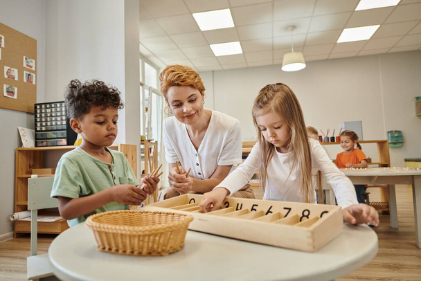 Montessori okulundaki derste çocuklar arasında gülümseyen öğretmen ellerinde tahta sopalarla. - Fotoğraf, Görsel