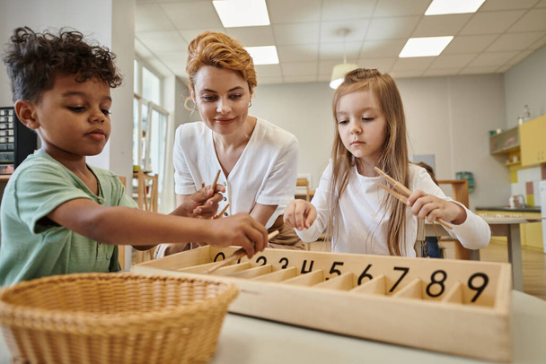 Montessori okulunda gülümseyen öğretmenin yanında tahta sopalarla oynayan çok ırklı çocuklar. - Fotoğraf, Görsel