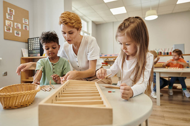 učitel mluví s multietnické děti při hraní s dřevěnými tyčinkami v Montessori škole - Fotografie, Obrázek