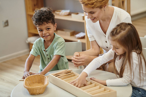 happy african american boy playing and looking at camera near teacher and friend in montessori class - Foto, Bild
