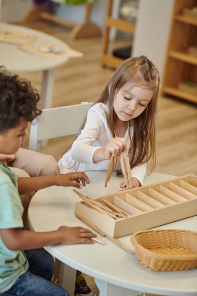 międzyrasowe dzieci bawiące się drewnianymi kijami na stole podczas lekcji w szkole montessori - Zdjęcie, obraz