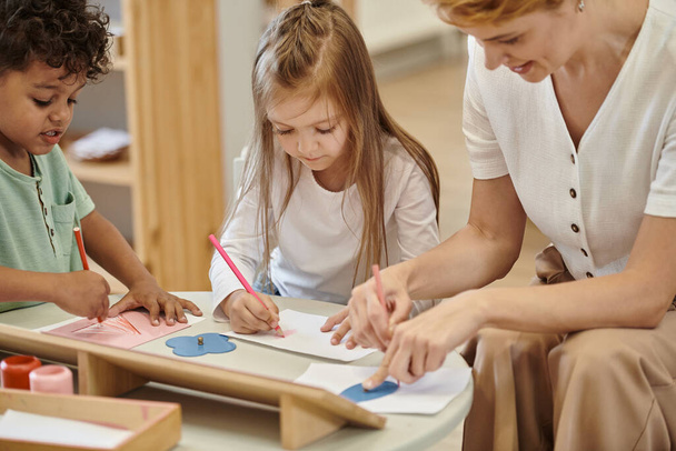 többnemzetiségű gyerekek rajzolnak ceruzával és beszélnek a mosolygós tanár közelében a montessori iskolában - Fotó, kép